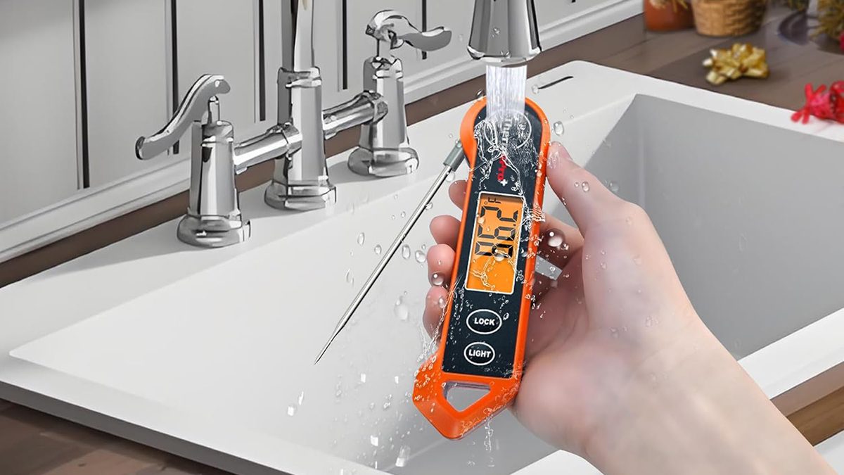 A hand holding an orange thermometer under running water