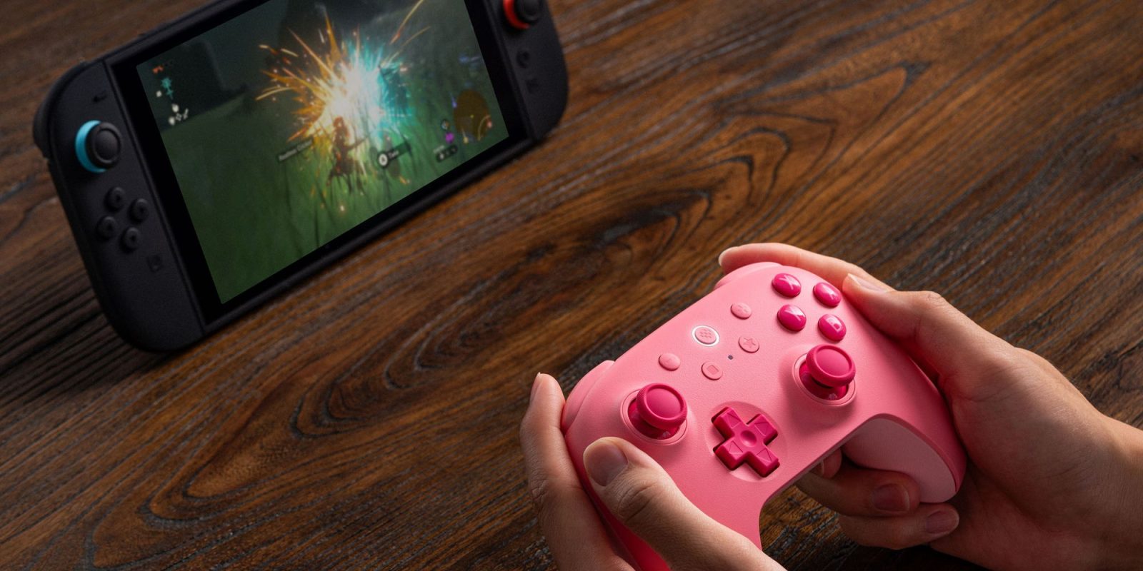 Image showing a person holding 8Bitdo's Ultimate 2C wireless controller in pink in front of the Switch 2.