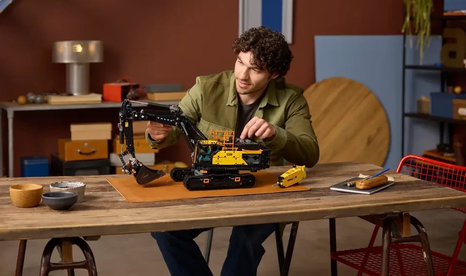 A man sitting at a table with a LEGO Technic Volvo EC500 Hybrid excavator set