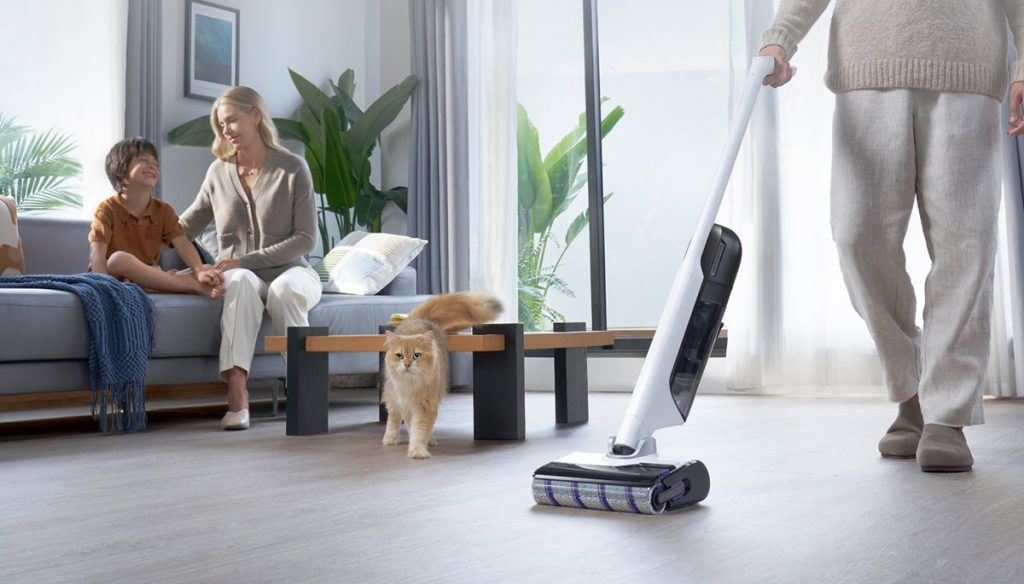 A person holding a vacuum cleaner with a cat on the floor