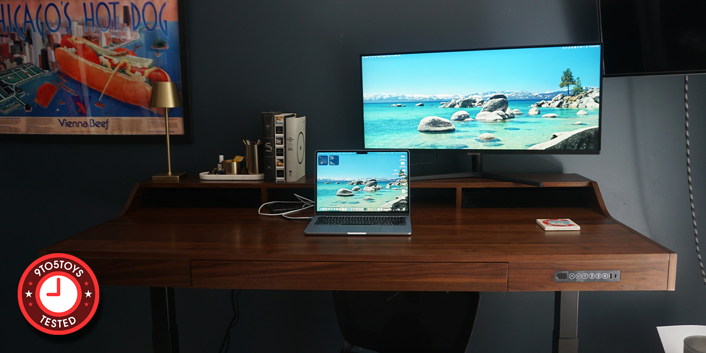 A laptop on a desk with a monitor and a picture on the wall