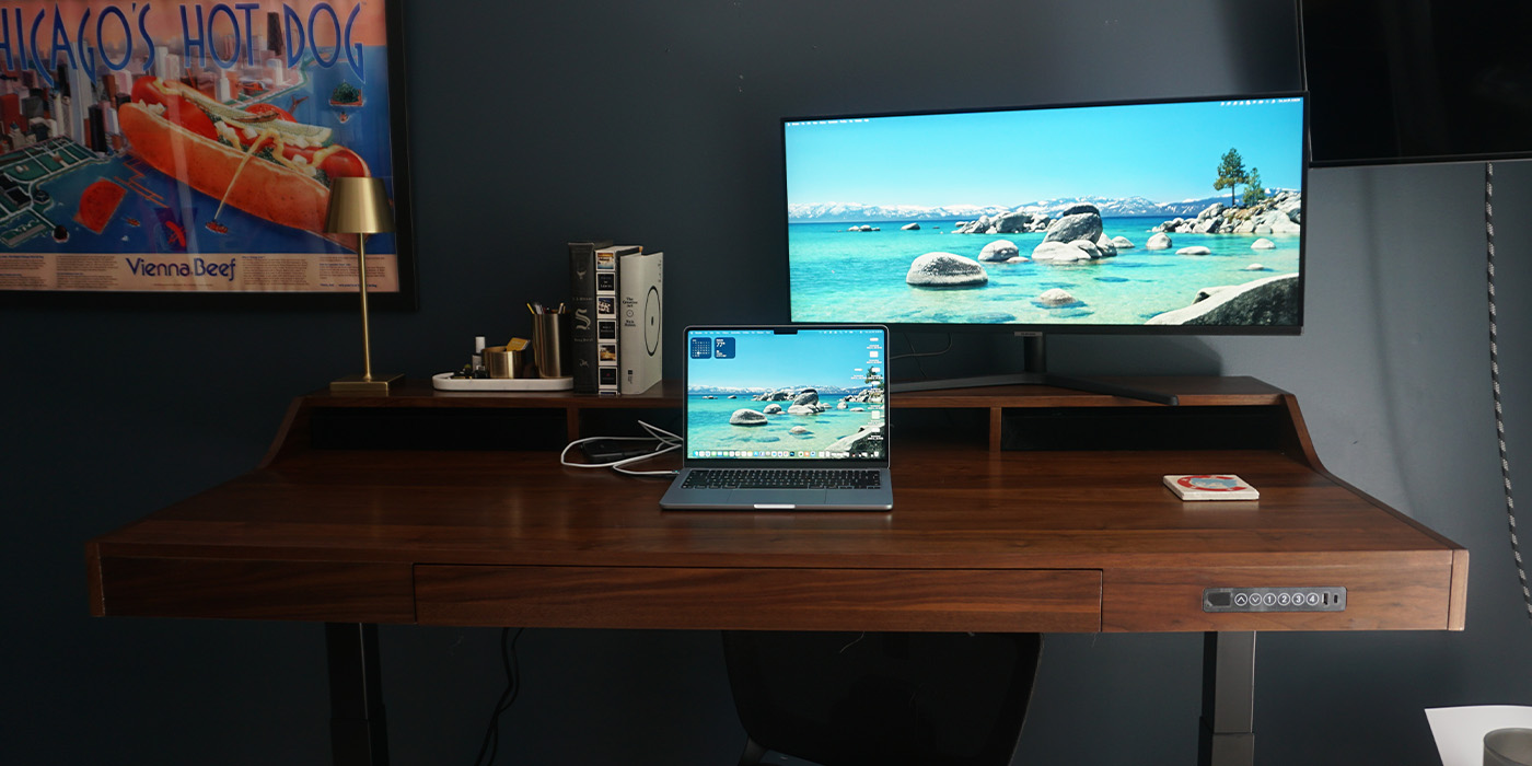 A laptop on a desk with a monitor and a picture on the wall